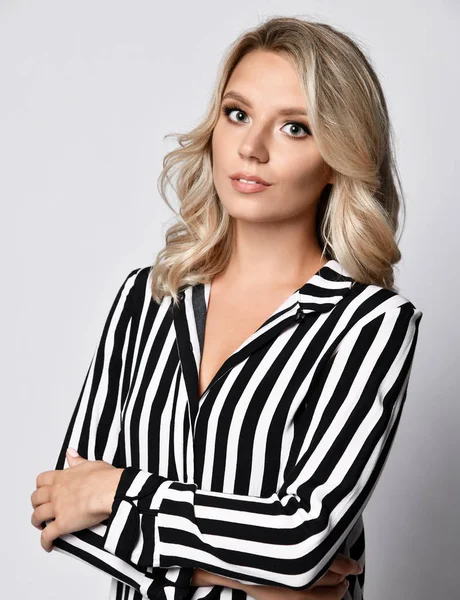 Puzzled looking young elegant business woman in formal wear striped blouse stands with her hands crossed