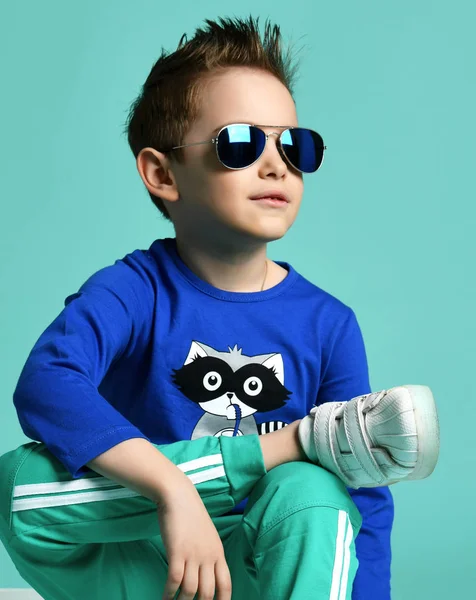 Cool młody chłopiec Kid w niebieskie okulary Aviator i niebieska koszula z nadrukiem Raccoon siedzi z nogami cool skrzyżowane — Zdjęcie stockowe