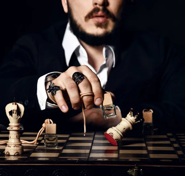 Brutal man in black suit and stylish rings playing chess with bottle of perfume, making a move, attack white queen