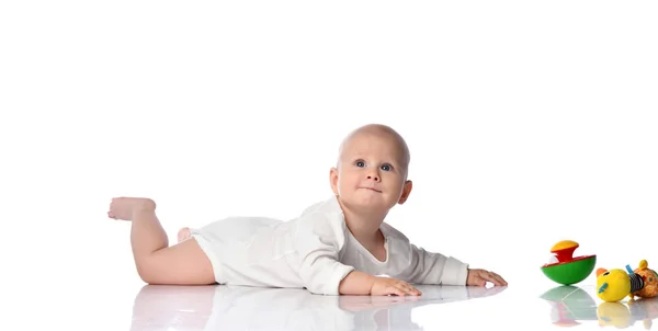 Baby kind baby jongen peuter in wit bodysuit liggend op zijn buik op zoek omhoog kruipen naar kleurrijke speelgoed te krijgen op wit — Stockfoto