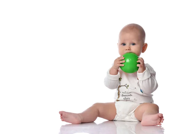 Младенец мальчик в подгузнике и белой рубашке сидит с зеленым шариком игрушки в руках, кусая его на белом — стоковое фото