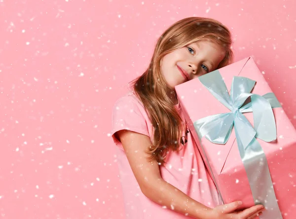 Weihnachts- und Neujahrskonzept. Lächelndes blondes Mädchen im rosafarbenen Kleid, das eine große Geschenkbox in der Hand hält und sich auf rosa umarmt — Stockfoto