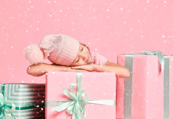 Koncept Vánoc a Nového roku. Malá holčička v pleteném klobouku spí na velkých dárkových krabicích pod sněhem na růžové — Stock fotografie