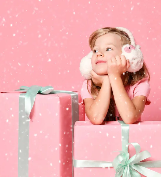 Christmas and New Year concept. Smiling blonde kid girl in fur earmuffs is sitting behind present gifts waiting on pink — ストック写真