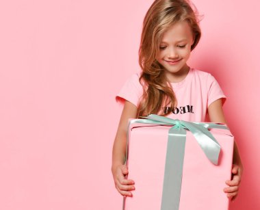 Joyful blonde kid girl in pink dress holding a big present gifts box looking at it impatiently on pink with copy space clipart