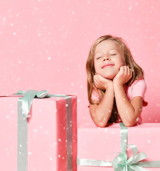 Noel et Nouvel An concept. Enfant fille est assis derrière des cadeaux avec son menton dans les mains sous la neige sur rose — Photo