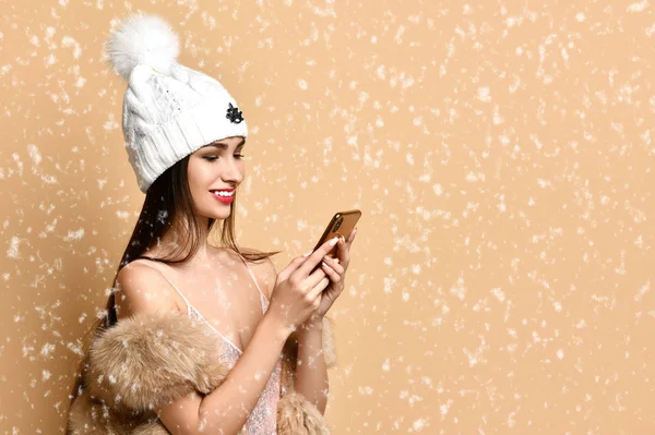 Joyful woman in party dress, fur and winter hat is looking at  her cell phone under the snow on beige with copy space — Stockfoto