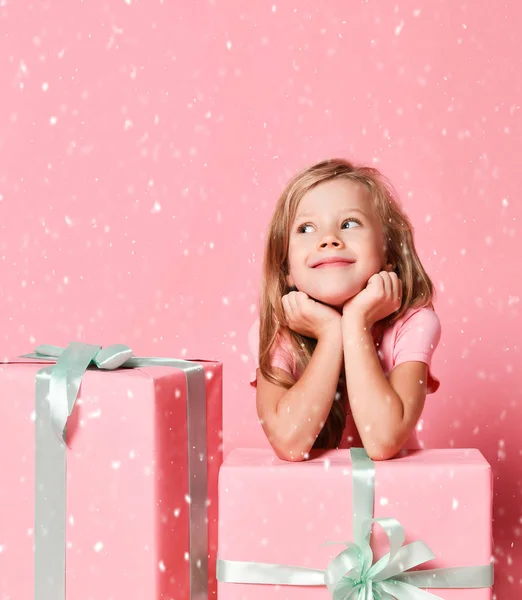 Christmas and New Year concept. Cute kid girl at gifts boxes with her chin in her hands dreaming under the snow on pink — Stock Photo, Image