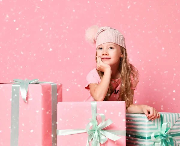 Enfant fille est assis à des boîtes-cadeaux en essayant de deviner ce qui est à l'intérieur, prêt à obtenir son présent sous la neige sur rose — Photo
