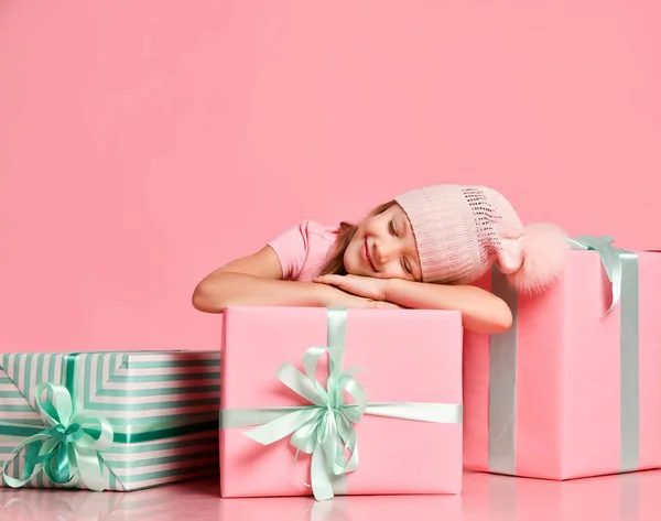 Natale e Capodanno concetto. Ragazzina sorridente in divertente cappello a maglia dorme su scatole regalo guardando i sogni su rosa — Foto Stock