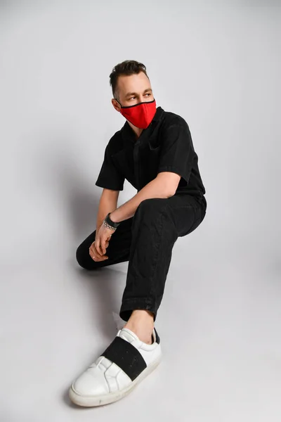黒い服と金属アクセサリーの若い男は感染から保護するために赤い医療用マスクで不法占拠座っている — ストック写真