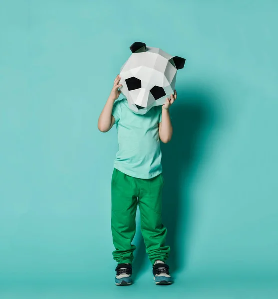 Panda maskeli, beyaz, boş tişörtlü, yeşil pantolonlu çocuk hayal kırıklığına uğramış gibi elleriyle başını tutuyor. — Stok fotoğraf