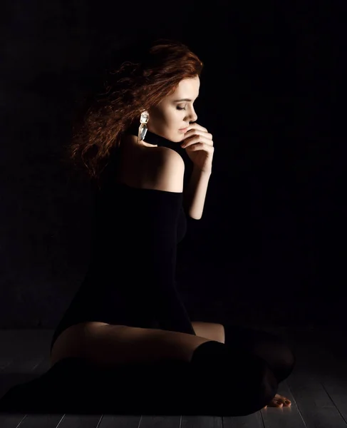 Mulher encaracolada jovem bonita em roupa interior do corpo, meias altas pretas e brincos sentados no chão, tocando o rosto e olhando para baixo — Fotografia de Stock