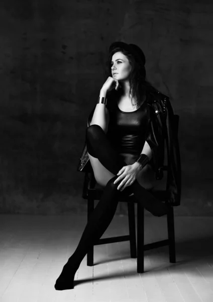 Mulher pensativa jovem em roupa interior do corpo, jaqueta de couro, meias altas, chapéu e acessórios senta-se na cadeira olhando para o canto superior — Fotografia de Stock