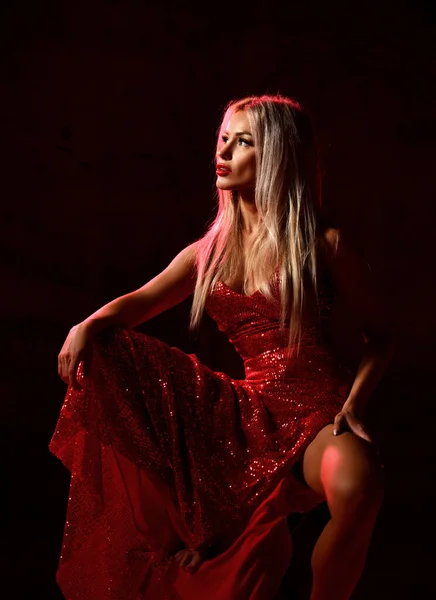 Mladá krásná blondýna žena v sexy červené lesklé šaty s hlubokým výstřihem sedí na židli a dívá se stranou — Stock fotografie