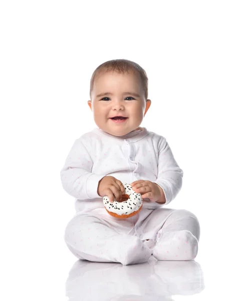 Happy laughing infant baby toddler in white onepiece overall sits on the floor holding donut in hands — Stock Photo, Image