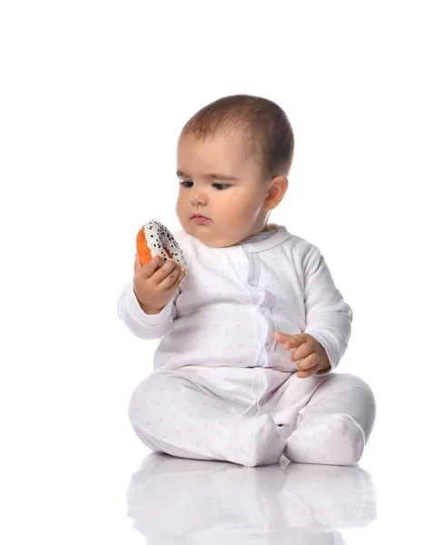 Thoughtful infant baby toddler in white onepiece overall sits on the floor looking at donut she holds in hands — Stock Photo, Image