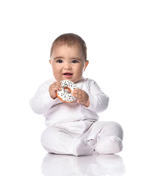 Happy laughing infant baby toddler in white onepiece overall sits on the floor holding donut in hands going to bite — Stock Photo, Image
