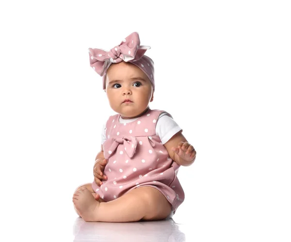 Focused concentrated infant baby girl toddler in polka-dot dress and headband with bow sits on the white floor — Stock Photo, Image