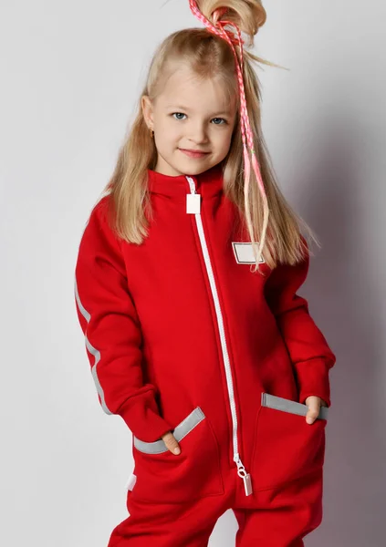 Portrait de jeune fille blonde froncée en combinaison rouge et coiffure cool debout avec ses mains dans les poches — Photo