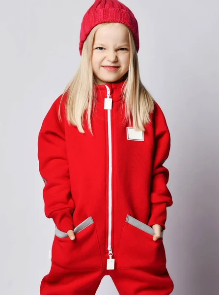 Ritratto di ragazza bionda maliziosa in tuta rossa e cappello invernale in piedi con le mani in tasca — Foto Stock