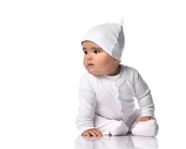 Curious infant baby toddler sits on the floor in white onepiece jumpsuit overall and funny hat with ears and looks aside — Stock Photo, Image