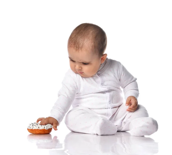 Цікавий дитячий малюк у білому комбінезоні для стрижки на лижах сидить на підлозі і підбирає пончик — стокове фото