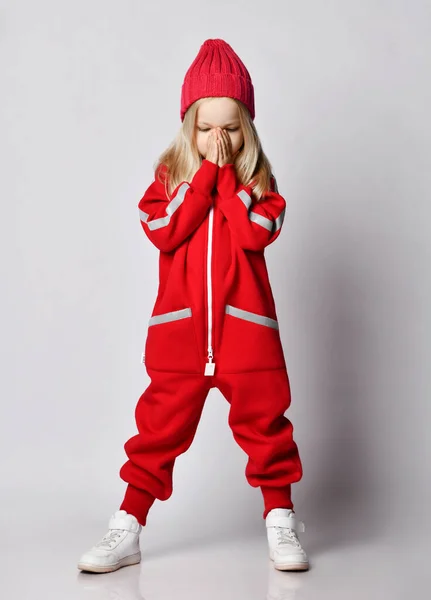 Blonde kid girl in red overall jumpsuit and winter hat stands holding hands at her nose covering — Stock Photo, Image