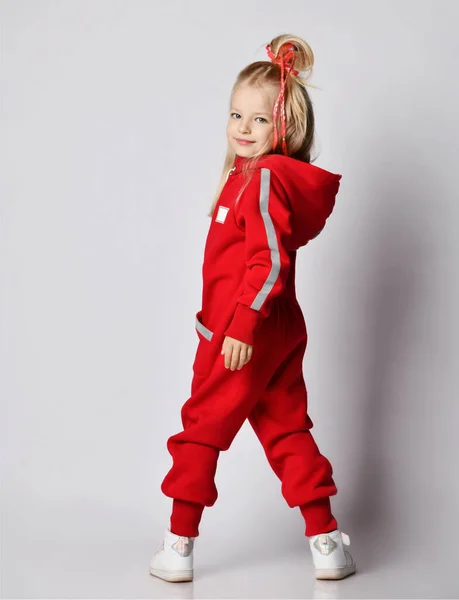 Blonde kid girl in red jumpsuit with hood and cool hairstyle stands back to us and turned. Back view. Full-growth. — Stock Photo, Image