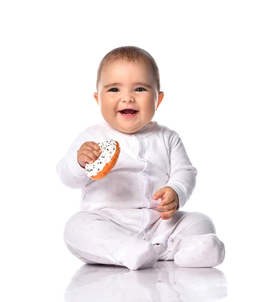 Gürültülü bir şekilde gülen, bembeyaz tulum giymiş bebek bebek, elinde donutla yerde oturuyor ve el sallıyor. — Stok fotoğraf