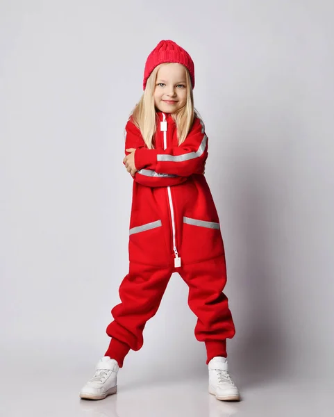 Smiling blonde kid girl in red overall jumpsuit and winter hat stands with her hands crossed at chest hugging herself — Stock Photo, Image