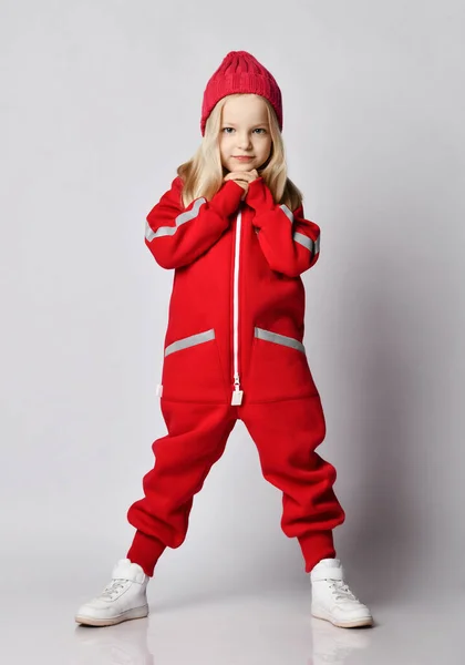 Full-growth portrait of blonde kid girl in red overall jumpsuit and winter hat standing with her hands at her chin — Stock Photo, Image