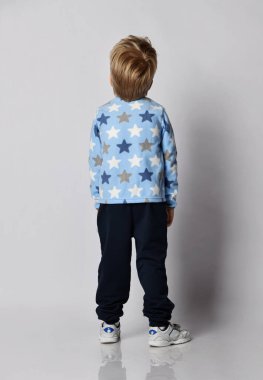 Mavi pantolonlu sarışın çocuk, yıldız desenli yün ceketli süveter ve beyaz spor ayakkabılar bize geri dönüyor.