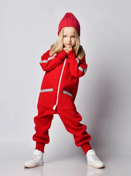Retrato de crescimento completo de menina loira em macacão geral vermelho e chapéu de inverno balançando com as mãos no queixo — Fotografia de Stock