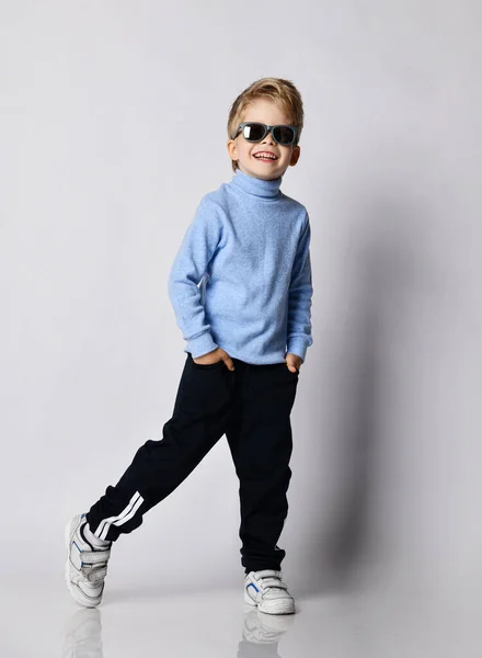 Niño rubio en zapatillas de deporte, pantalones deportivos negros, jersey de cuello alto azul y gafas de sol con el pie a un lado —  Fotos de Stock