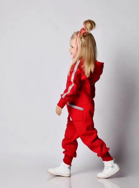 Stylish blonde kid girl in red coldproof overall jumpsuit and cool hairstyle passing us by, walking, leaving room — Stock Photo, Image