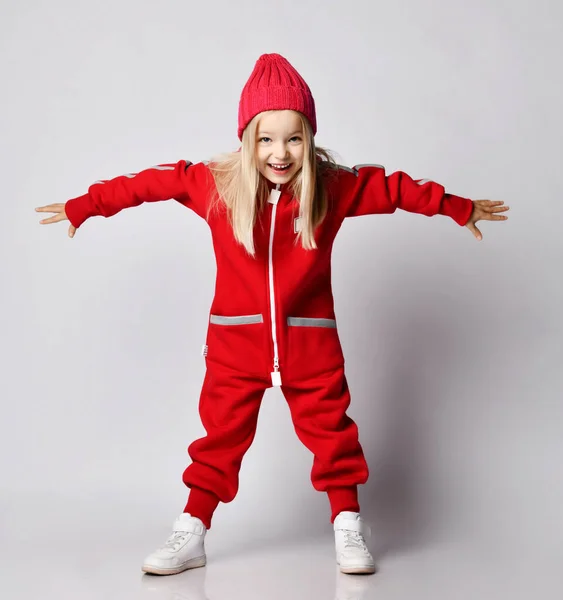 Playful blonde kid girl in red overall jumpsuit and winter hat standing with her arms spread wide like wings — Stock Photo, Image