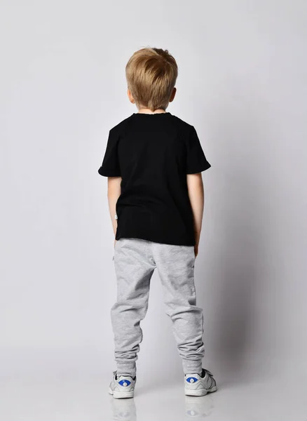 Menino loiro de t-shirt preta, calça cinza e tênis volta para nós com as mãos nos bolsos — Fotografia de Stock