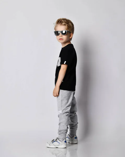 Ξανθό παιδί με μαύρο t-shirt, γκρι παντελόνι και sneakers στέκεται δίπλα μας με το κεφάλι γυρισμένο και κοιτάζει την κάμερα — Φωτογραφία Αρχείου