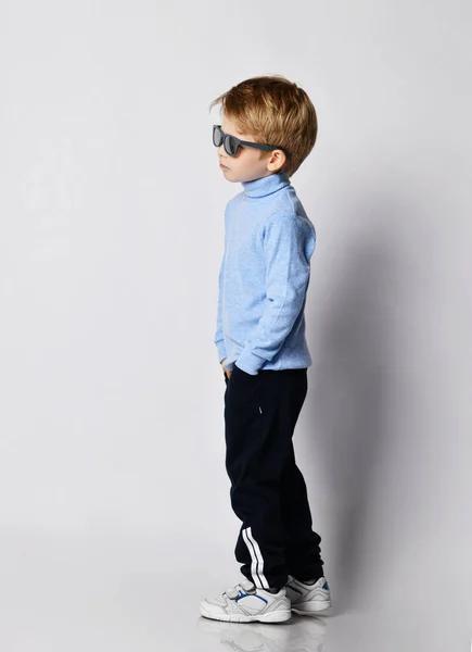 Sebevědomý blonďatý chlapec v teniskách, černých sportovních kalhotách, modrém roláku svetru a slunečních brýlích stojí při nás — Stock fotografie