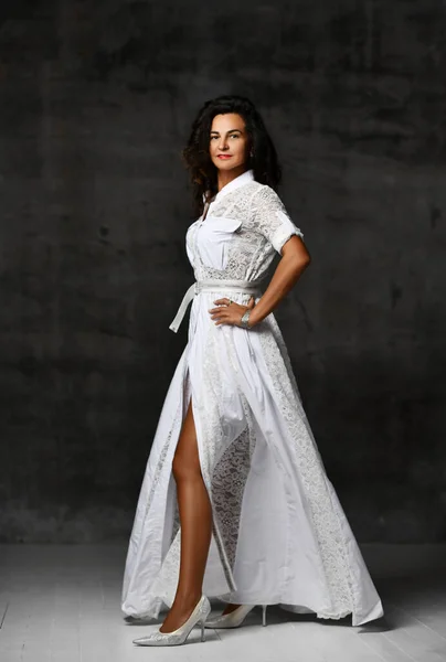 Elegante kraftvolle brünette Frau in weißem langen Spitzenkleid mit kurzen Ärmeln und hochhackigen Schuhen steht vor der Kamera — Stockfoto