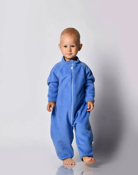 Positieve blote voeten blond baby jongen in blauwe fleece jumpsuit met rits staat houden voet omhoog — Stockfoto