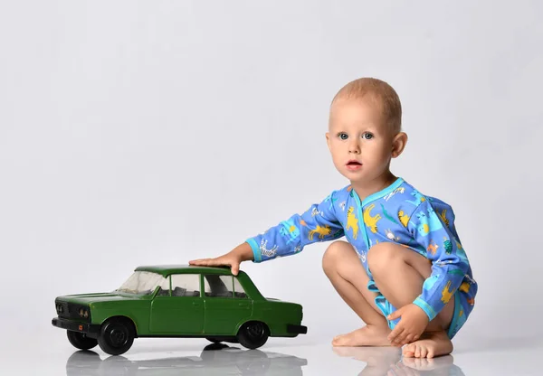 Descalzo bebé niño en azul de una sola pieza del traje de mameluco con mangas largas se sienta en cuclillas juega con el coche de juguete grande —  Fotos de Stock