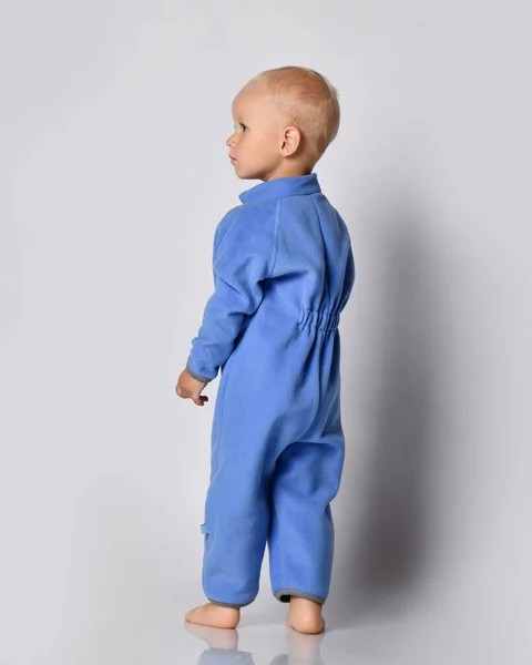 Volle groei portret van blote voeten baby jongen in blauwe fleece jumpsuit staan terug naar camera kijken opzij — Stockfoto