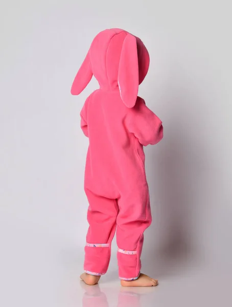 분홍색 따뜻하고 편안 한 점프 복을 입고 후드를 쓰고 토끼 귀는 카메라 앞에 뒤로 서 있는 발가벗은 아기 — 스톡 사진