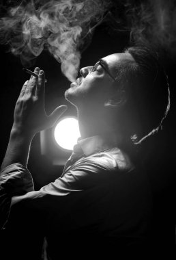 Resmi tişörtlü ve gözlüklü bir adamın portresi. Kafası yukarıda sigara içiyor, gözleri kapalı ve üflüyor. Yan görünüm