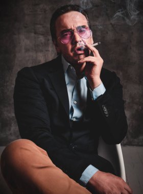 Şık resmi takım elbiseli ve füme mavi gözlüklü, sakallı, sakallı vahşi bir adam sigara içiyor.