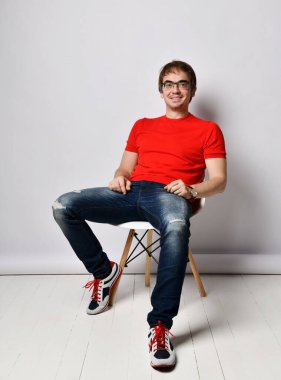 Kırmızı tişörtlü, mavi kotlu, spor ayakkabılı ve gözlüklü gülümseyen orta yaşlı bir adam rahat bir şekilde sandalyede oturuyor.