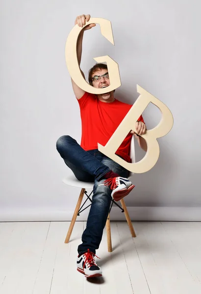 Hombre de mediana edad en camiseta roja, vaqueros azules, zapatillas de deporte y gafas se sienta en la silla con grandes letras G y B y lo muerde —  Fotos de Stock