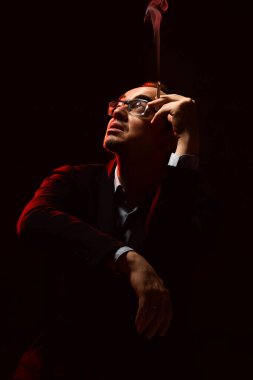 Resmi takım elbiseli ve gözlüklü orta yaşlı bir adam sandalyeye oturup sigara içiyor ve spot ışıklarına bakıyor.